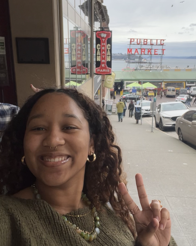 Kymari making a peace sign with the Pike Place Market sign behind her in Seattle